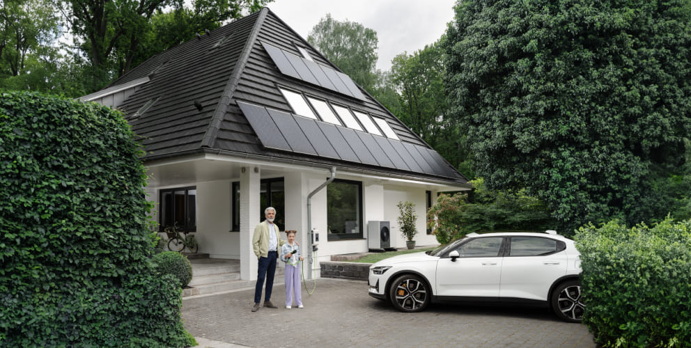 Ein Mann steht neben einem Kind vor einem Einfamilienhaus mit Solarpanelen auf dem Dach. Neben ihnen parkt ein Elektroauto in der Einfahrt.