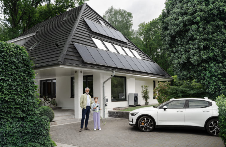 Ein Mann steht neben einem Kind vor einem Einfamilienhaus mit Solarpanelen auf dem Dach. Neben ihnen parkt ein Elektroauto in der Einfahrt.