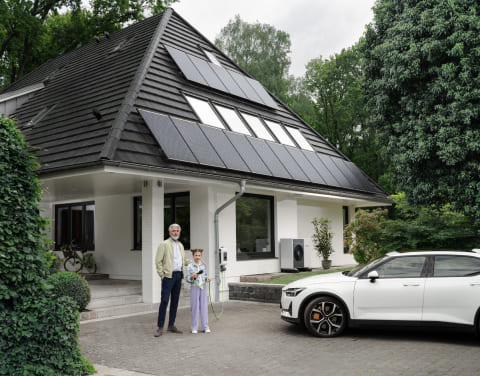 Ein Mann steht neben einem Kind vor einem Einfamilienhaus mit Solarpanelen auf dem Dach. Neben ihnen parkt ein Elektroauto in der Einfahrt.