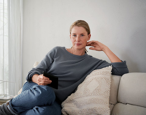 Eine Frau sitzt entspannt auf einem Sofa und schaut in die Kamera.