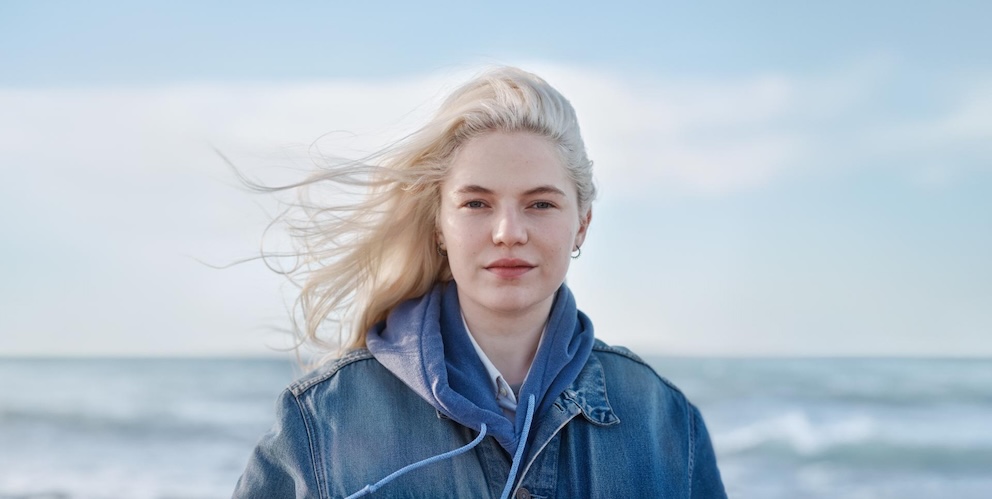 Frau steht vor einem Meer und die Haare wehen im Wind.