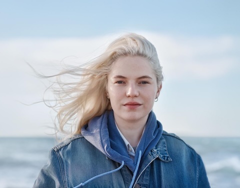 Frau steht vor einem Meer und die Haare wehen im Wind.