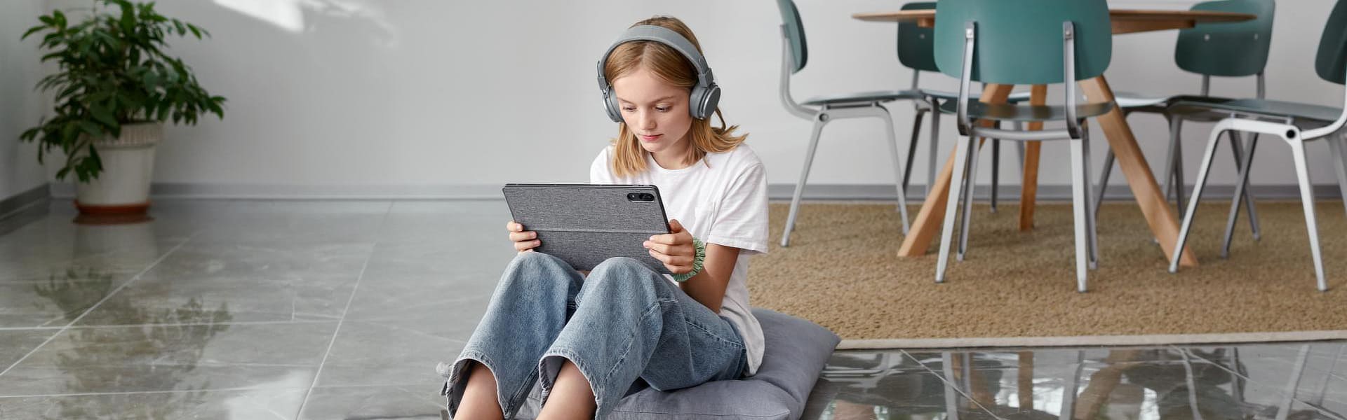 Mädchen sitzt im Wohnzimmer auf einem Fliesenfußboden und schaut auf ein Tablet.