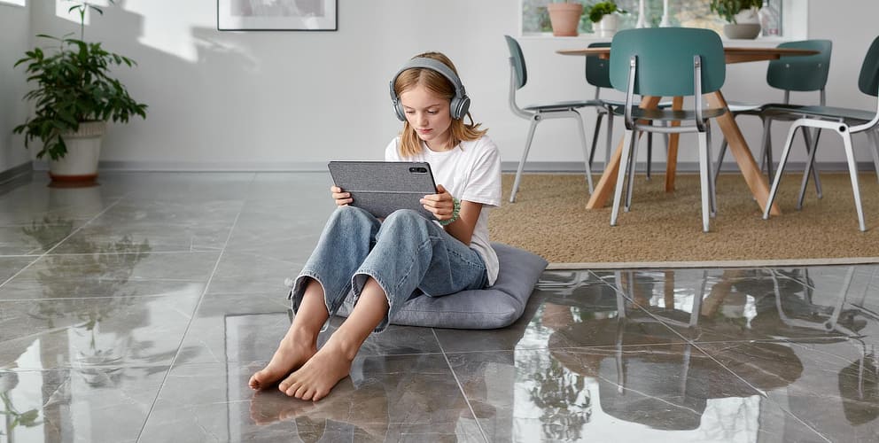 Mädchen sitzt im Wohnzimmer auf einem Fliesenfußboden und schaut auf ein Tablet.
