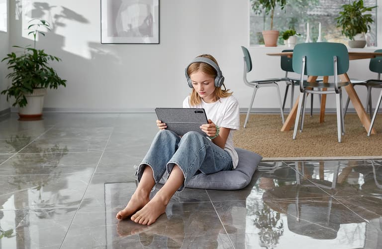 Mädchen sitzt im Wohnzimmer auf einem Fliesenfußboden und schaut auf ein Tablet.