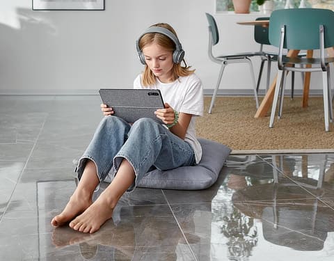 Mädchen sitzt im Wohnzimmer auf einem Fliesenfußboden und schaut auf ein Tablet.
