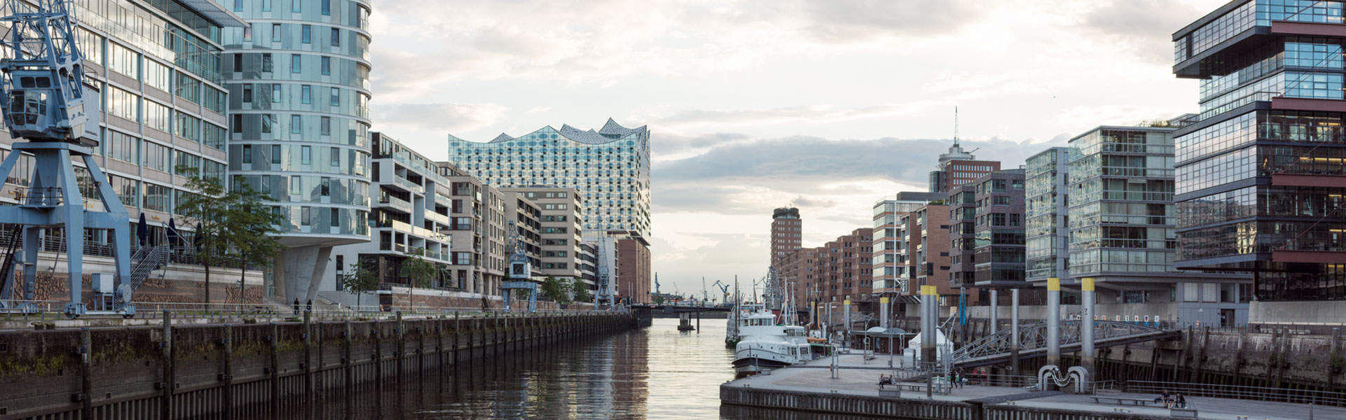 Aufnahme der Stadt Hamburg vom Wasser aus, mit Blick auf die Elbphilharmonie. Im Wasser liegen vereinzelt Schiffe. Links und rechts vom Hafen liegen Neubauwohnungen der HafenCity.