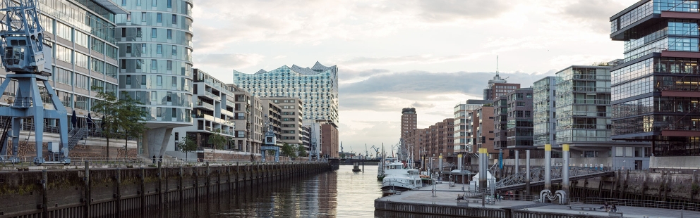 Aufnahme der Stadt Hamburg vom Wasser aus, mit Blick auf die Elbphilharmonie. Im Wasser liegen vereinzelt Schiffe. Links und rechts vom Hafen liegen Neubauwohnungen der HafenCity.