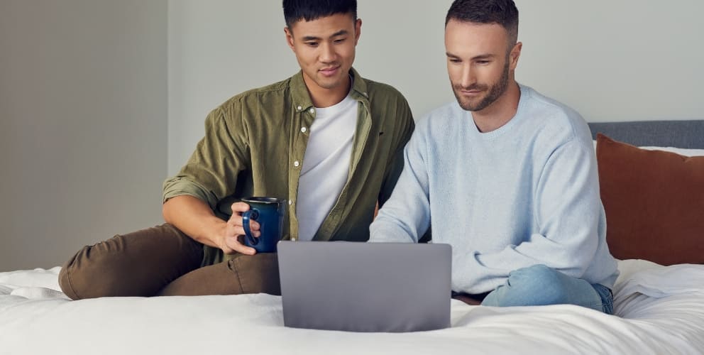 Zwei Männer sitzen entspannt auf einem Bett und schauen auf einen Laptop.