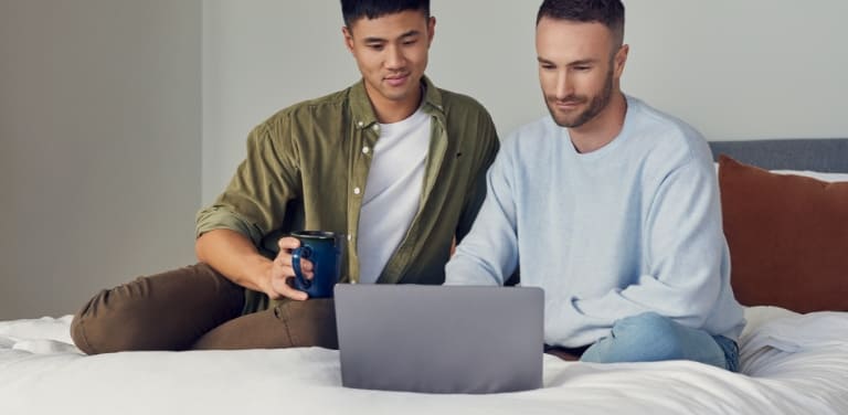 Zwei Männer sitzen entspannt auf einem Bett und schauen auf einen Laptop.