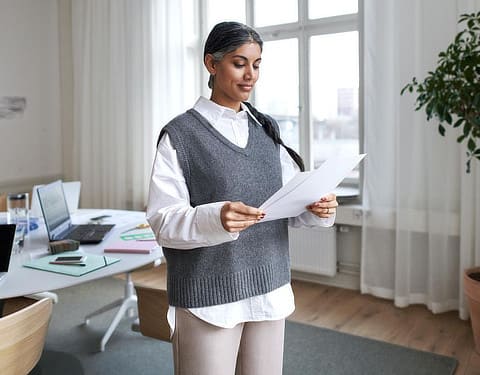 Eine Frau steht in einem hellen und modernen Arbeitszimmer. Sie hält mehrere Zettel in der Hand und liest diese aufmerksam durch.