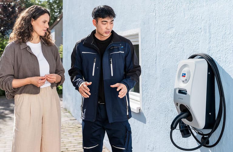 Kundin und Fachpersonal führen Einweisungsgespräch neben einer Wallbox an einer Hausfassade.