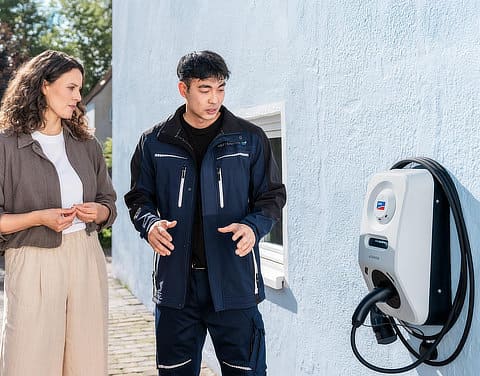 Kundin und Fachpersonal führen Einweisungsgespräch neben einer Wallbox an einer Hausfassade.