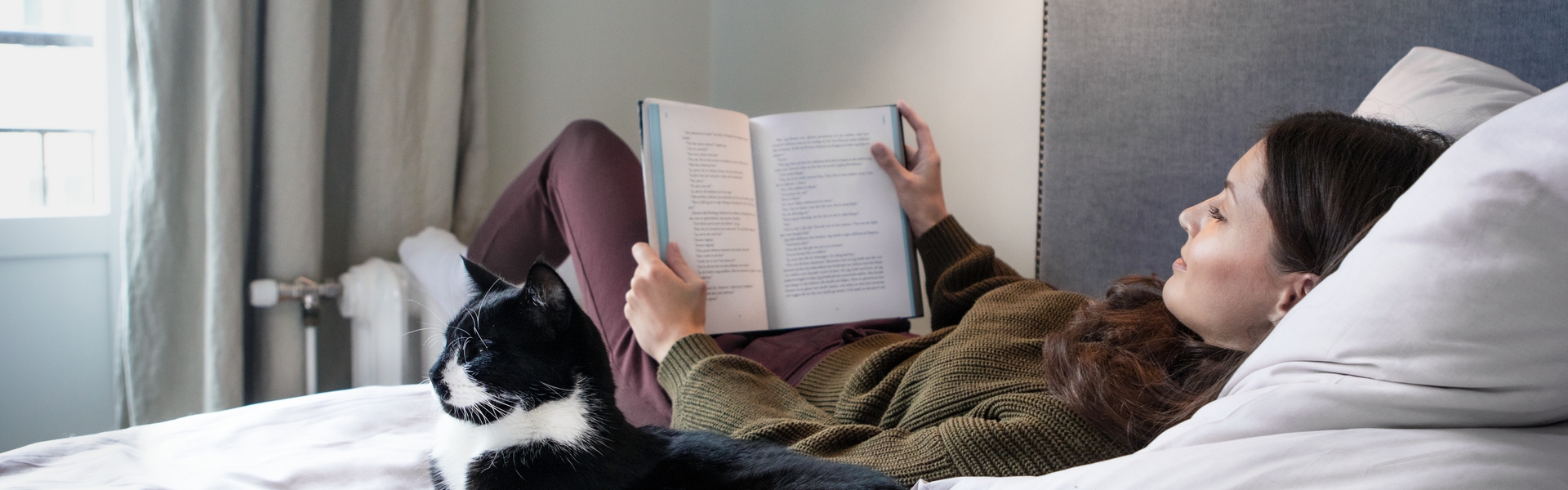 Frau liegt lesend auf einem Bett neben einer Katze. Ihre Füße wärmt die Frau an einem Heizkörper. Beide befinden sich in einem modernen und hellen Schlafzimmer.