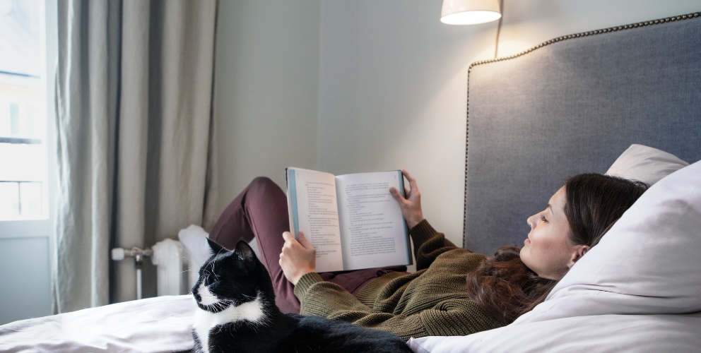 Frau liegt lesend auf einem Bett neben einer Katze. Ihre Füße wärmt die Frau an einem Heizkörper. Beide befinden sich in einem modernen und hellen Schlafzimmer.
