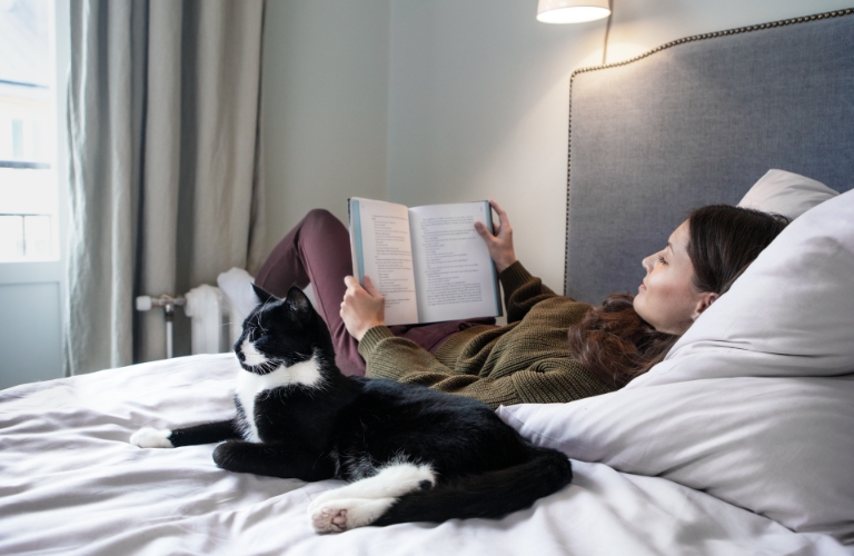 Frau liegt lesend auf einem Bett neben einer Katze. Ihre Füße wärmt die Frau an einem Heizkörper. Beide befinden sich in einem modernen und hellen Schlafzimmer.
