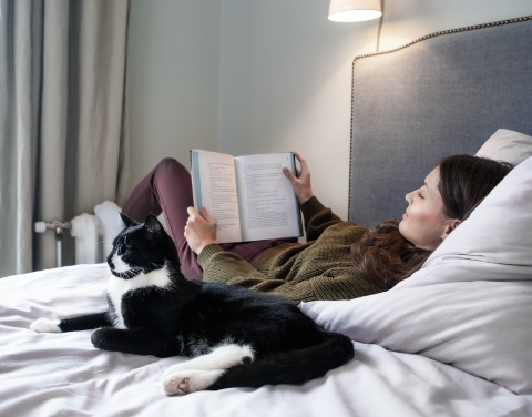 Frau liegt lesend auf einem Bett neben einer Katze. Ihre Füße wärmt die Frau an einem Heizkörper. Beide befinden sich in einem modernen und hellen Schlafzimmer.