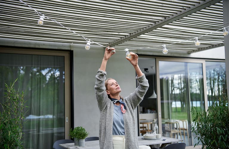 Frau steht draußen auf einer Terrasse. Sie hält die Glühbirne einer Außenlichterlichterkette in der Hand und betrachtet sie.