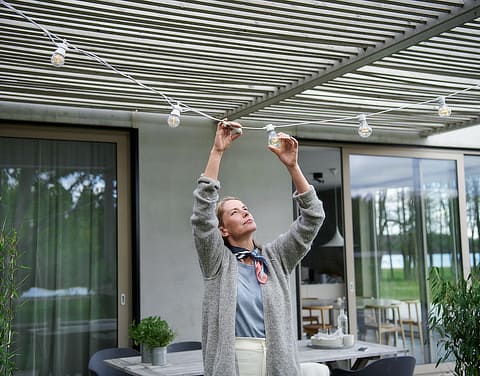Frau steht draußen auf einer Terrasse. Sie hält die Glühbirne einer Außenlichterlichterkette in der Hand und betrachtet sie.