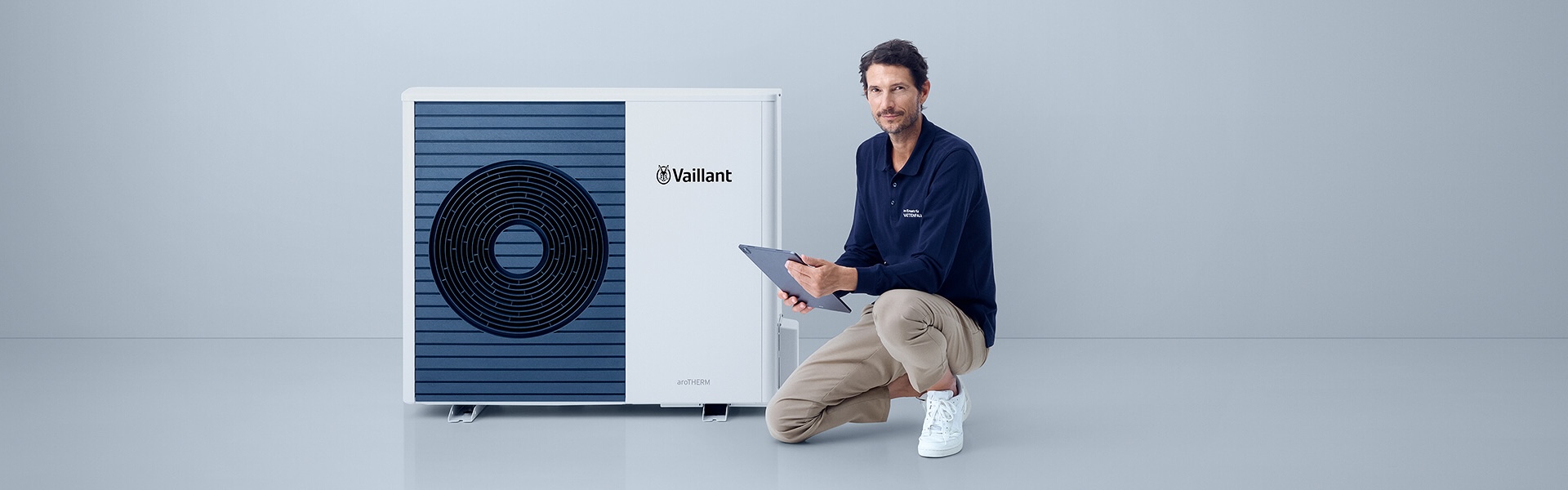 Ein locker gekleideter Mann kniet mit einem Bein neben einer modernen Wärmepumpe. Er hält ein Tablet in der Hand. Der Hintergrund ist schlicht und hell.