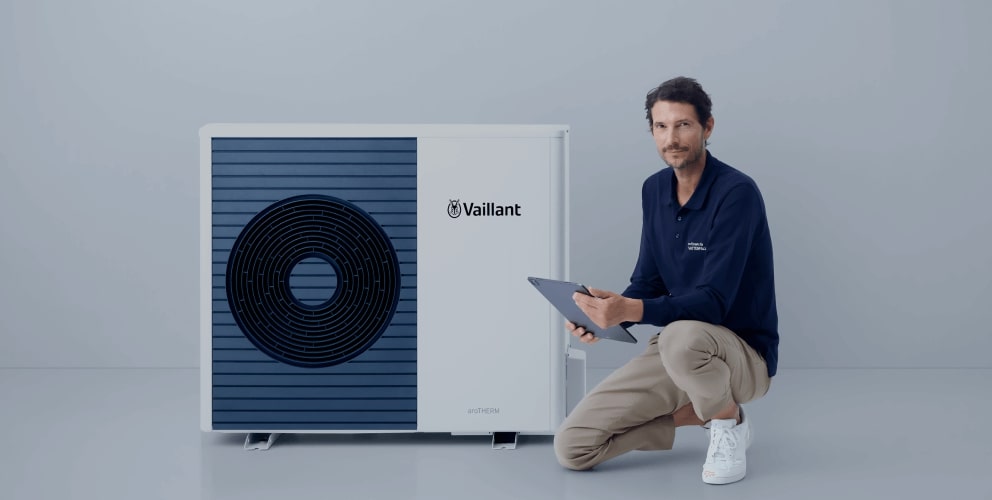 Ein locker gekleideter Mann kniet mit einem Bein neben einer modernen Wärmepumpe. Er hält ein Tablet in der Hand. Der Hintergrund ist schlicht und hell.