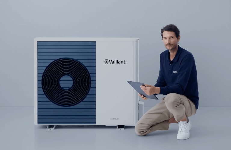 Ein locker gekleideter Mann kniet mit einem Bein neben einer modernen Wärmepumpe. Er hält ein Tablet in der Hand. Der Hintergrund ist schlicht und hell.