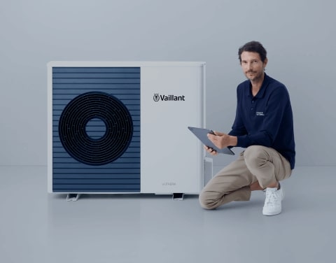 Ein locker gekleideter Mann kniet mit einem Bein neben einer modernen Wärmepumpe. Er hält ein Tablet in der Hand. Der Hintergrund ist schlicht und hell.