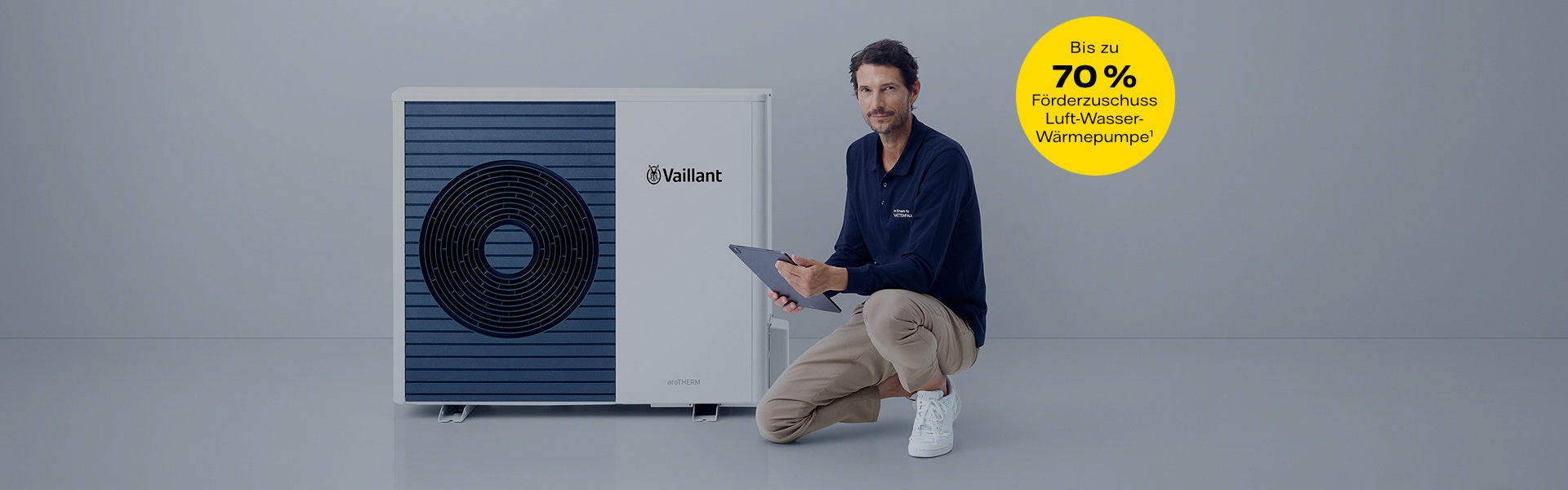 Ein locker gekleideter Mann kniet mit einem Bein neben einer modernen Wärmepumpe. Er hält ein Tablet in der Hand. Der Hintergrund ist schlicht und hell.