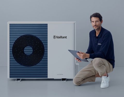 Ein locker gekleideter Mann kniet mit einem Bein neben einer modernen Wärmepumpe. Er hält ein Tablet in der Hand. Der Hintergrund ist schlicht und hell.